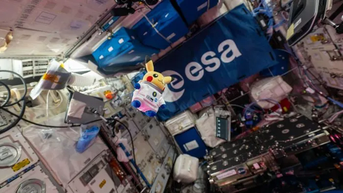 The Astronaut Pikachu plush floating aboard the ISS in front of an ESA flag